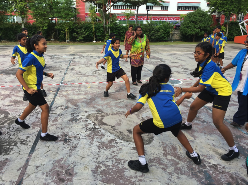 Children in blue and yellow uniforms play a sport on an outdoor court while an adult watches nearby.